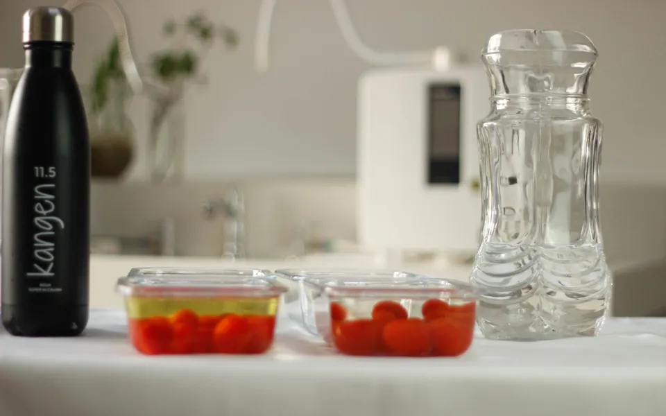 Garrafa Kangen Água Super Alcalina ao lado de potes de vidro com tomates-cereja e uma jarra de água, sobre uma mesa branca